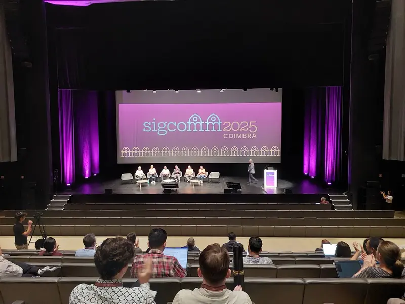 People seated on the SIGCOMM 2025 stage in front of a full auditorium