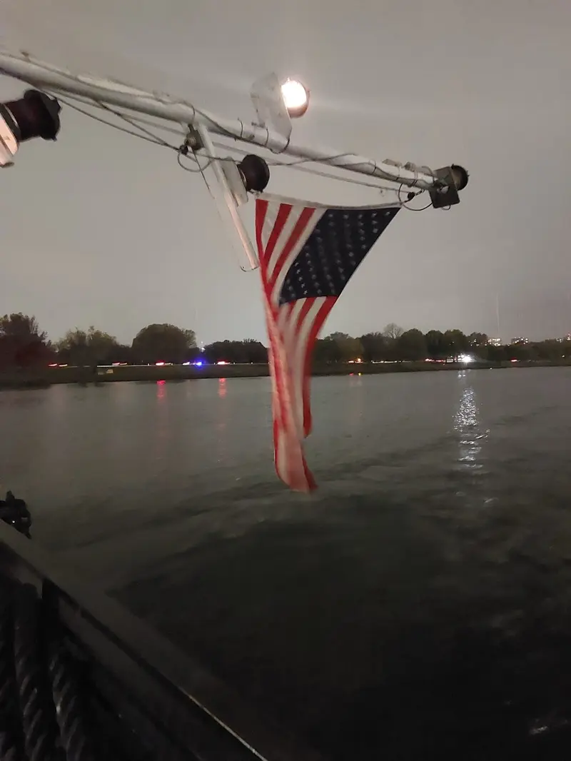 An American flag at the stern of the boat, the river and the shore lights at dusk